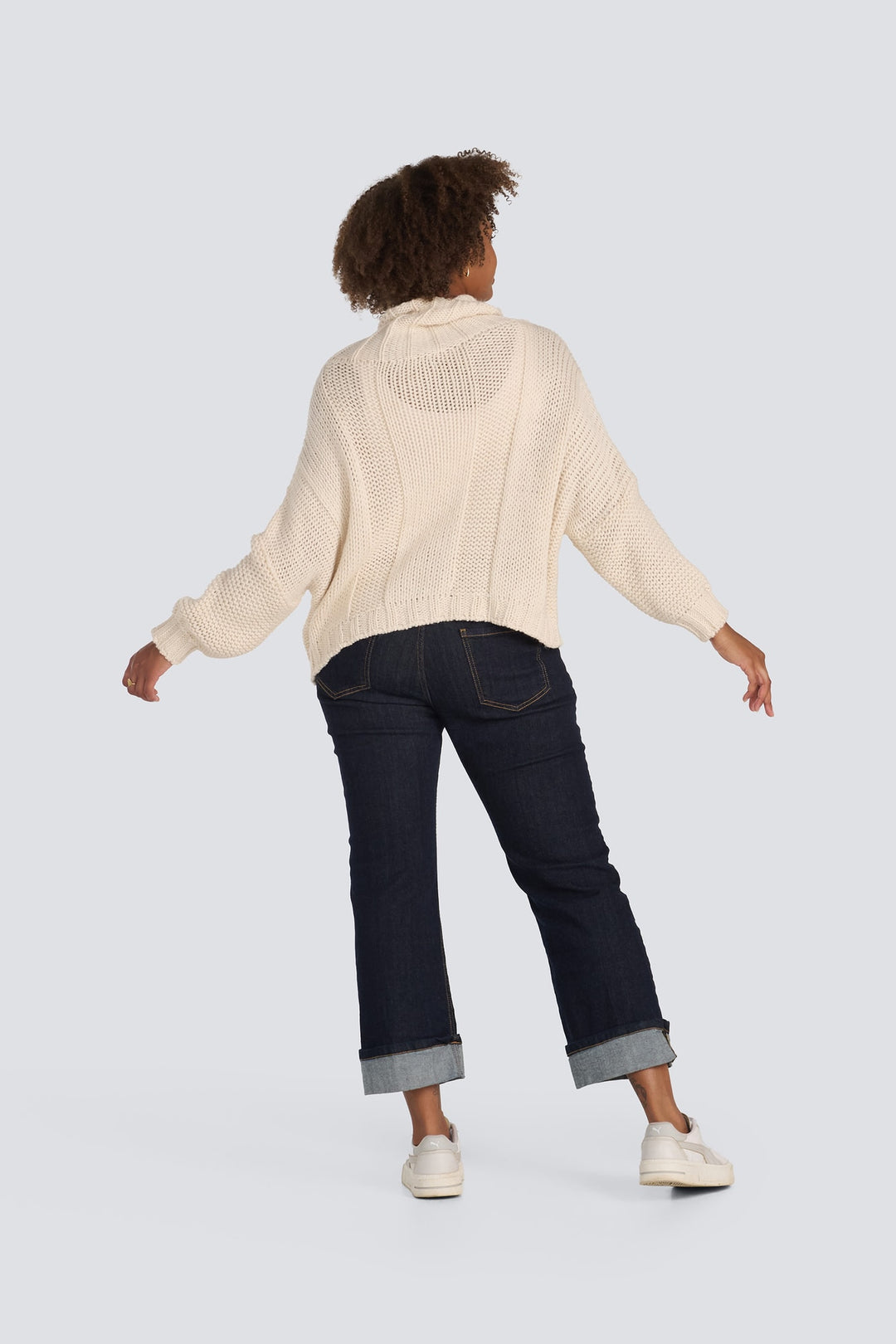 Chunky cream knit turtleneck sweater shown from behind with dark cuffed jeans.